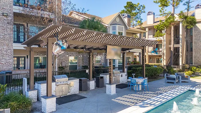 Inviting apartment community with a stylish outdoor grilling area, shaded pergola, and tranquil swimming pool under sunny skies.