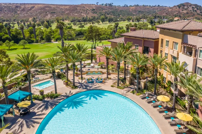 Elegant apartment complex with a resort-style pool, lush palm trees, and vibrant poolside seating, offering serene views of expansive green golf courses and rolling hills.