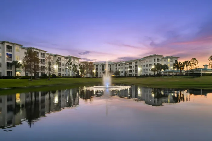 Elegant apartment complex with tranquil pond and fountain at sunset, surrounded by lush greenery.
