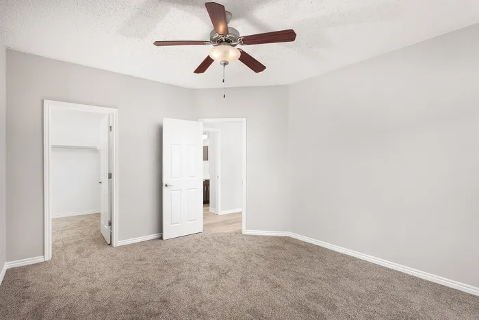 Bright and spacious bedroom with soft carpeting and modern ceiling fan, featuring white doors leading to a walk-in closet and a sunlit hallway.