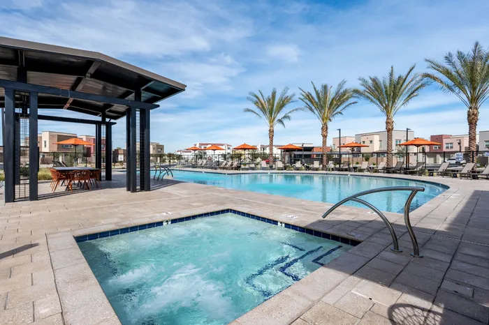 Luxurious apartment complex with sparkling pool, relaxing spa, shaded seating, and lush palm trees under a clear blue sky.