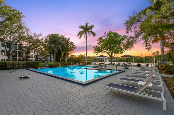 Inviting poolside area with lounge chairs, surrounded by lush greenery and modern apartments against a colorful sunset sky.