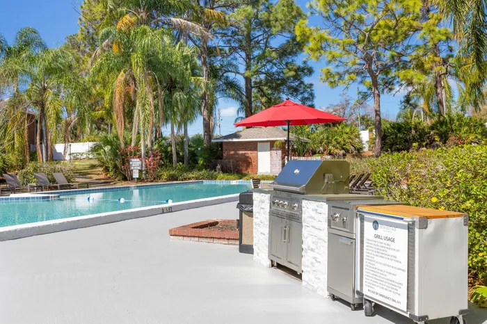 Beautiful poolside oasis with palm trees, outdoor grill station under a vibrant red umbrella, and comfortable lounge chairs for relaxation.