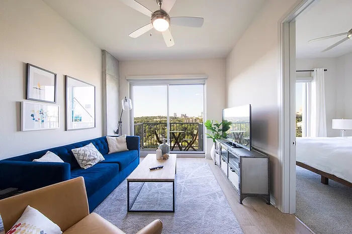 Bright modern living room with blue sofa, stylish decor, and balcony view, seamlessly connected to an elegant bedroom.