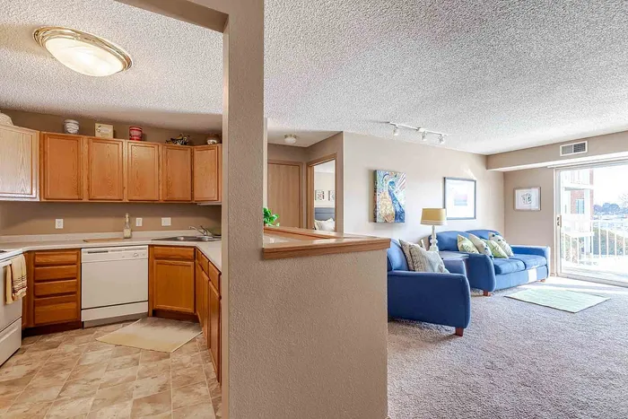 Bright open-concept living and kitchen area with cozy blue sofas, natural light, wooden cabinetry, and modern decor.