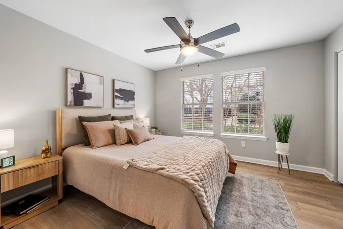 Cozy bedroom with a stylish bed, modern ceiling fan, and large windows letting in natural light, complemented by chic artwork and hardwood floors.
