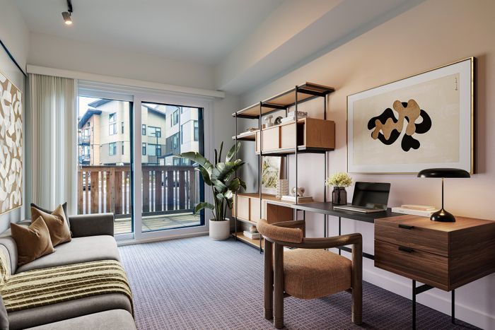 Bright and modern living space with a cozy sofa, stylish desk setup, elegant shelving, and large glass doors leading to a balcony with building views.