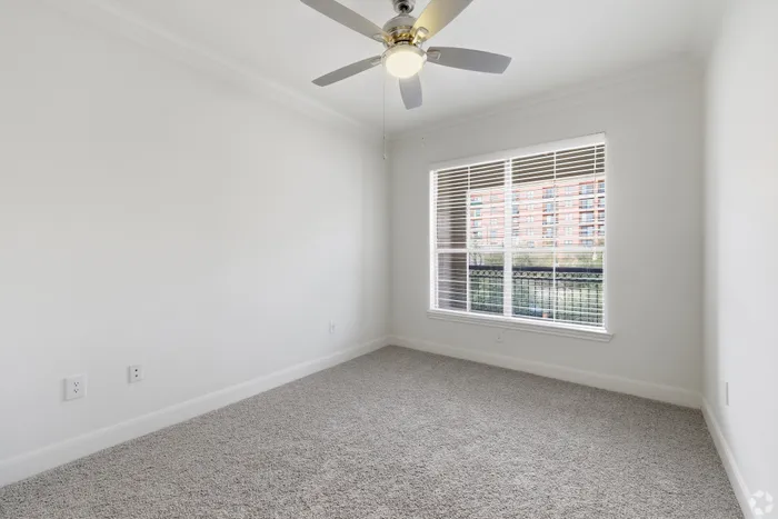 Bright apartment bedroom with plush carpet, large window offering city views, and modern ceiling fan.