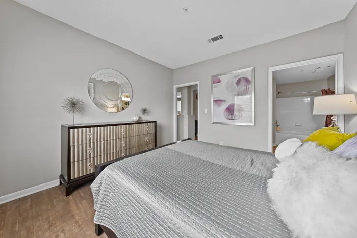 Elegant bedroom with modern decor featuring a plush bed, stylish dresser with unique mirror, and vibrant artwork, opening into a pristine bathroom.