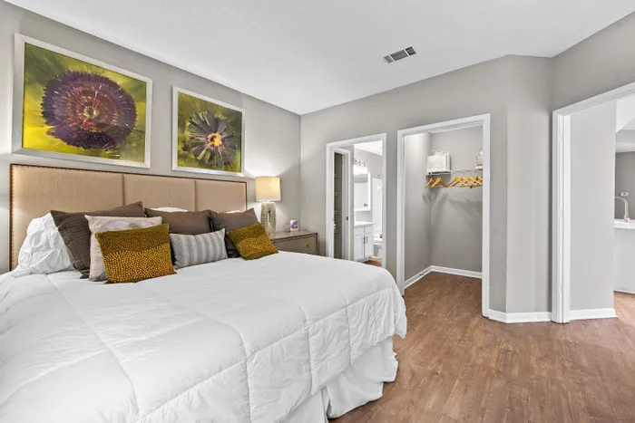 Elegant bedroom featuring a large cozy bed adorned with stylish cushions, modern artwork on walls, and rich wood flooring leading to a walk-in closet and ensuite bathroom.