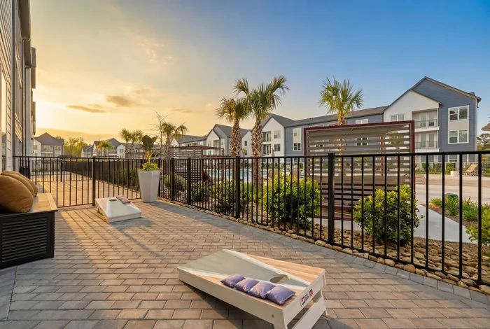 Inviting apartment patio with cozy seating, cornhole game, and scenic view of palm trees and modern buildings at sunset.