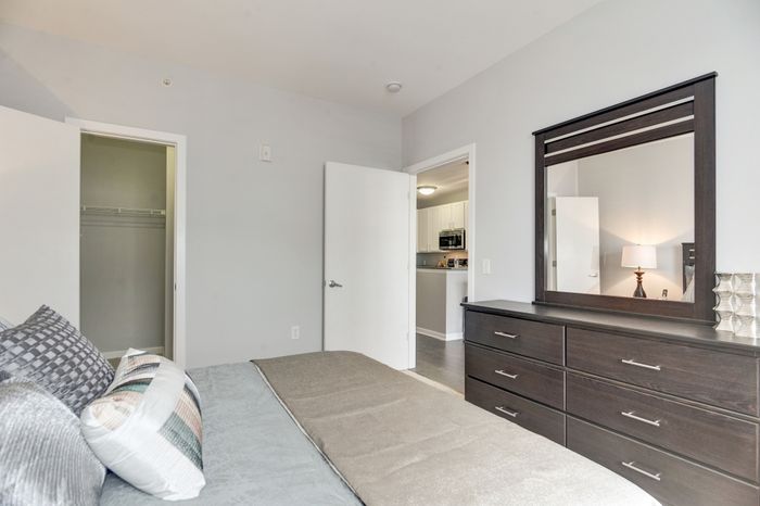 Modern bedroom with sleek dark wood dresser, large mirror, and cozy bed accented with stylish pillows, adjacent to an open doorway revealing a contemporary kitchen.