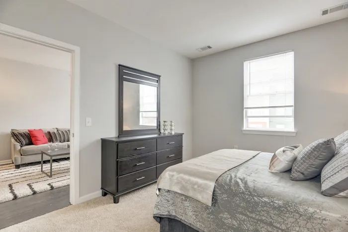 Bright and inviting bedroom with plush carpet, modern dresser, and window view, complemented by a cozy glimpse of the living area with stylish sofa.