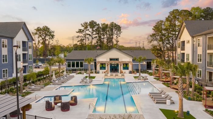A modern apartment complex courtyard featuring a large resort-style swimming pool with lounge chairs and umbrellas, surrounded by landscaped greenery and multi-story residential buildings, with a clubhouse in the background under a soft sunset sky.