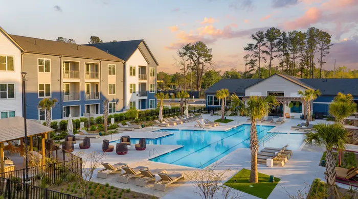A modern apartment complex courtyard featuring a large resort-style swimming pool with lounge chairs and umbrellas, surrounded by landscaped greenery and multi-story residential buildings, with a clubhouse in the background under a soft sunset sky.
