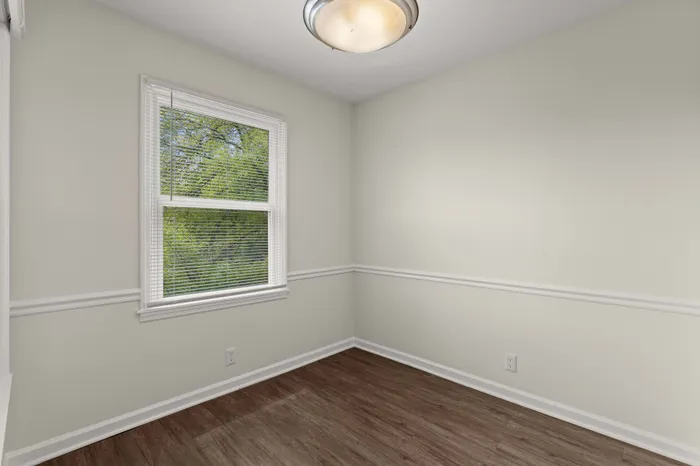 Bright and inviting room with chic wood flooring, fresh white walls, and a large window overlooking lush greenery, perfect for an office or cozy retreat.