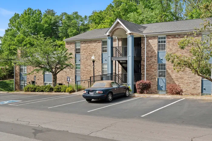 Charming brick apartment building surrounded by lush greenery, featuring ample parking and inviting entrance.