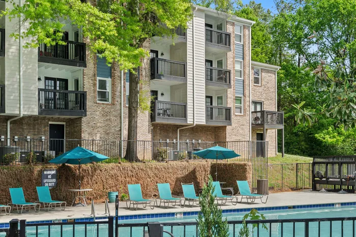 Inviting apartment complex with lush greenery, featuring modern balconies overlooking a sparkling pool adorned with teal loungers and umbrellas.