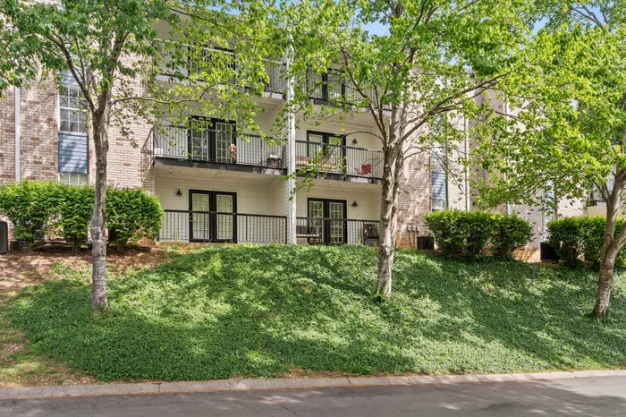 Charming apartment building with multiple patios, nestled in lush greenery and surrounded by mature trees, offering serene and scenic living.