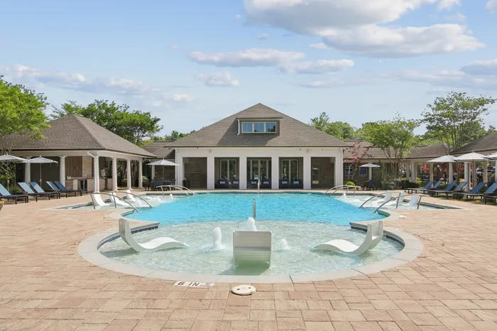Luxurious outdoor pool area with stylish lounge chairs and a modern clubhouse, surrounded by lush greenery and under a clear blue sky.