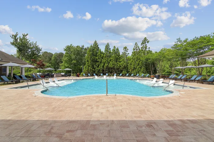Luxurious outdoor pool with sun loungers and umbrellas, surrounded by lush greenery under a bright blue sky.