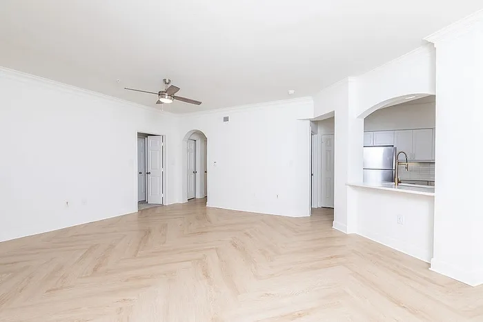 Bright open living space with elegant light wood flooring, ceiling fan, and modern kitchen featuring stainless steel appliances.