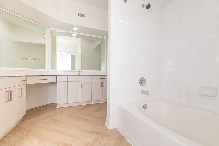 Bright and spacious bathroom featuring a large mirror, elegant vanity cabinets, and a pristine bathtub with white tile surround.