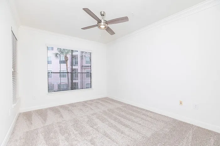 Bright, spacious bedroom with clean carpet, large window, and modern ceiling fan in a stylish apartment.