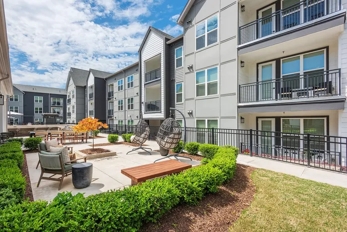 Elegant apartment complex featuring modern architecture with spacious balconies, lush garden seating area, and stylish hanging chairs under a clear blue sky.