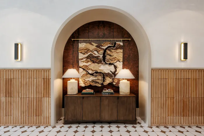 Elegant interior design featuring a stylish console with twin lamps, artistic woven tapestry, and warm wood accents under a graceful arch.
