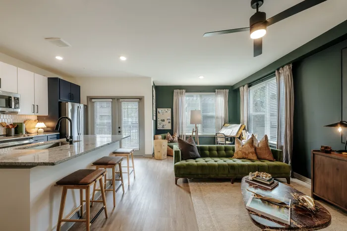 Modern open-concept living area with stylish kitchen, granite countertops, and cozy green sofa. Abundant natural light creates an inviting atmosphere.