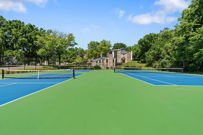 Vibrant outdoor tennis court surrounded by lush greenery and modern apartment buildings under a clear blue sky.