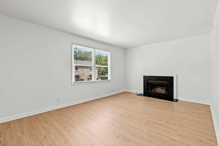 Bright and airy living room with sleek wood flooring, large window, and elegant fireplace, perfect for cozy evenings.