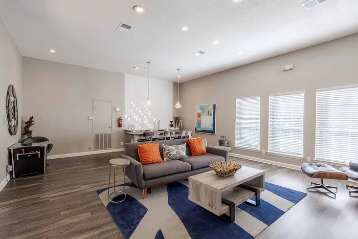 Chic living room with modern decor, featuring a cozy gray sofa with vibrant orange cushions, stylish coffee table, and elegant dining area under pendant lighting.