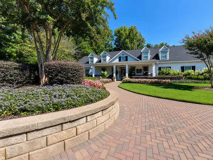 Charming white suburban house with perfectly manicured gardens and elegant brick pathway, nestled among lush greenery and vibrant flower beds.
