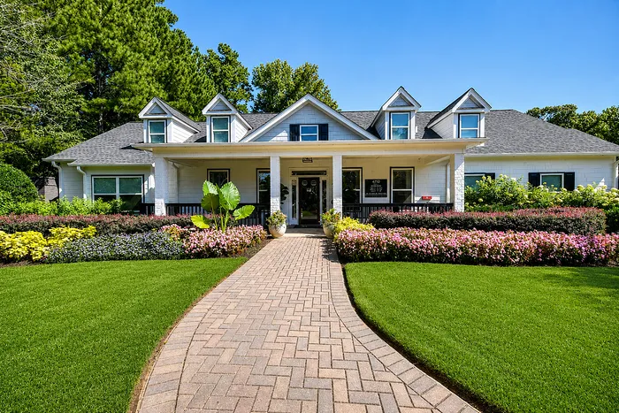 Charming white house with triangular dormers, surrounded by lush landscaping and a vibrant flower garden, featuring a welcoming brick pathway.