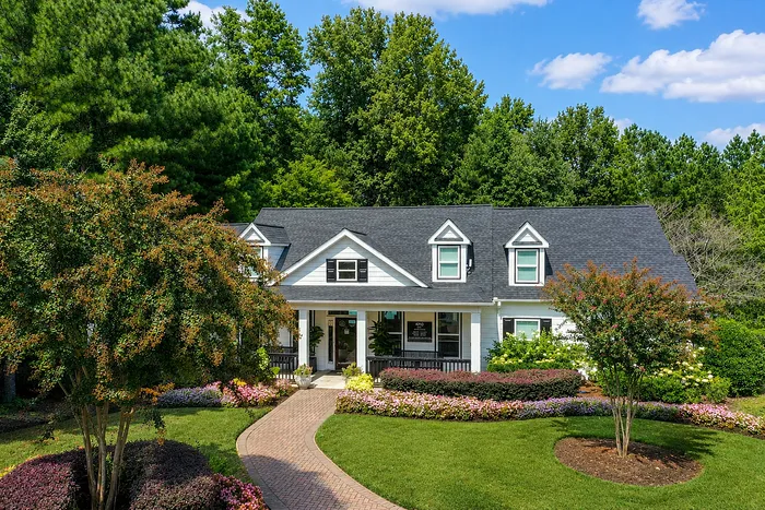 Charming colonial-style house with dormer windows, surrounded by lush greenery and vibrant flower beds on a sunny day.