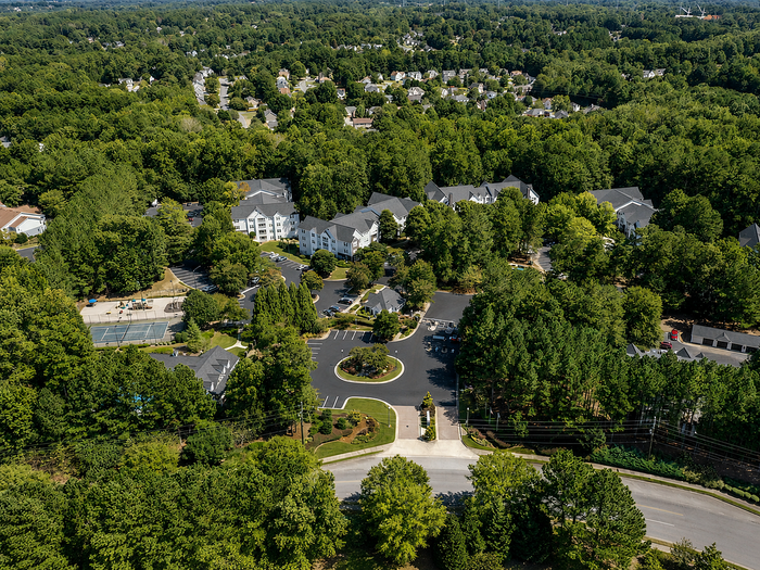 Scenic aerial view of a lush, green residential community with modern apartments, tennis courts, and ample parking, surrounded by vibrant trees.