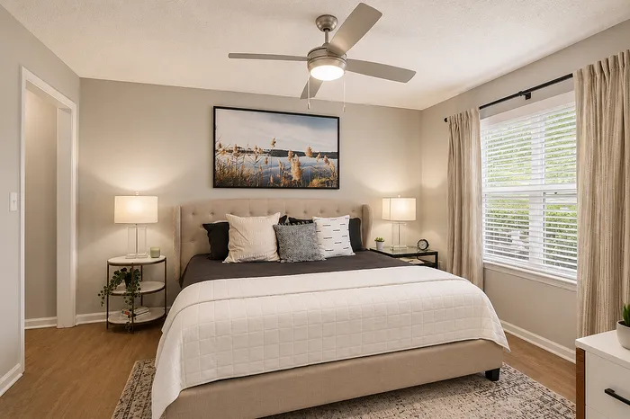Inviting bedroom with elegant neutral decor, featuring a plush bed with modern pillows, stylish lamps, serene wall art, ceiling fan, and natural light from a large window.