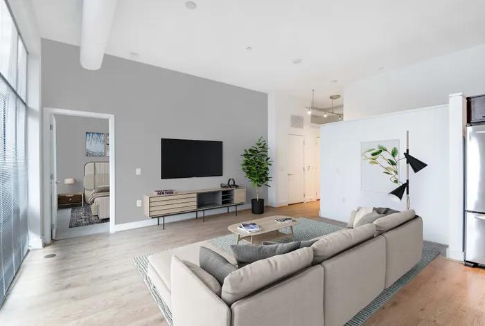Contemporary apartment living room with sleek decor, cozy sofa, wall-mounted TV, and bright natural light from large windows, leading to a stylish bedroom.