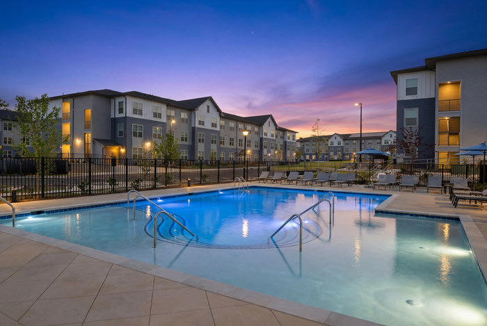 Resort-style swimming pool at dusk surrounded by modern apartment buildings, lounge seating, and illuminated community amenities.