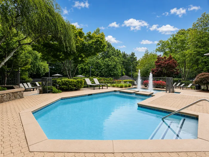 Elegant outdoor pool area surrounded by lush greenery, featuring modern sun loungers and a decorative fountain under a bright blue sky.
