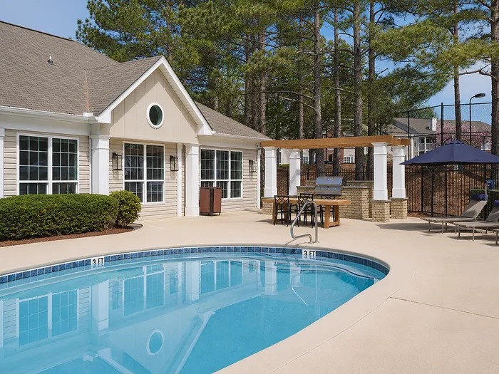 Charming outdoor pool area with sparkling water, elegant pergola, and built-in grill surrounded by lush greenery and cozy lounging chairs.