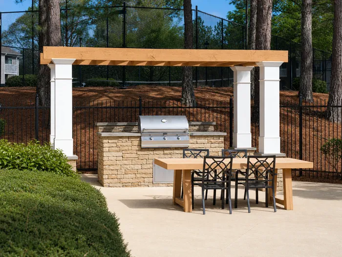 Charming outdoor barbecue area with a stone grill setup under a stylish pergola, featuring a wooden dining table and black chairs, nestled in a serene garden setting.