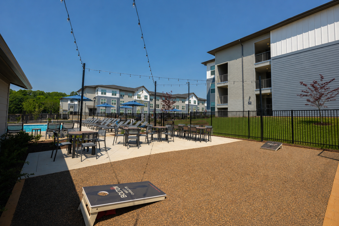 Modern apartment complex with inviting outdoor recreation area, featuring a sparkling pool, lounge chairs, tables with umbrellas, and a lawn game setup.