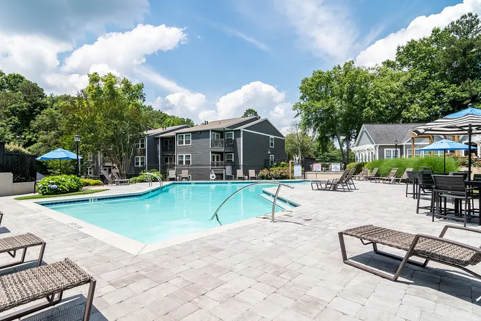 Inviting apartment complex with a sparkling pool, lounge chairs, and lush greenery under a bright blue sky.