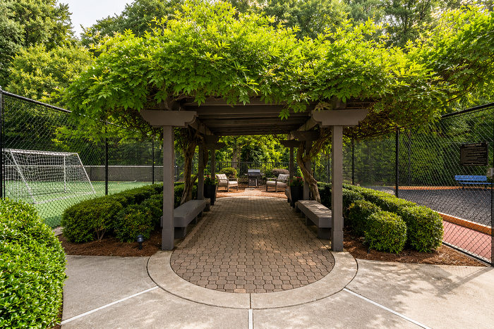 Charming outdoor pergola nestled in greenery, with comfortable seating and a view of a nearby soccer field.