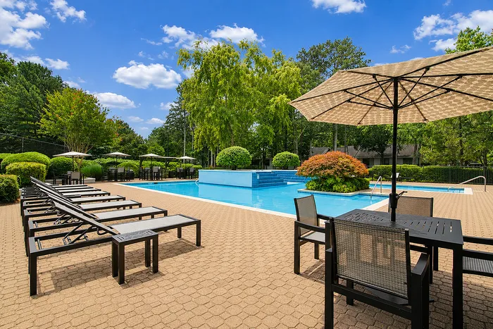 Luxurious outdoor pool area featuring modern lounge chairs, elegant patio seating, and lush greenery under a vibrant blue sky.