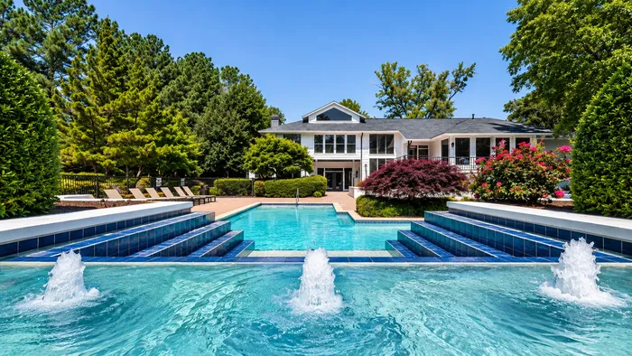 Luxurious poolside view of a modern white residence surrounded by lush greenery, featuring cascading fountains and comfortable sun loungers, offering a serene oasis retreat.