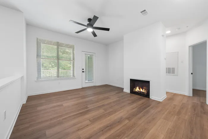 Bright and modern living room featuring elegant wood flooring, a cozy fireplace, and a ceiling fan, with natural light streaming through large windows.
